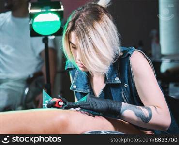 View of professional blonde tattoo female master is painting with black ink, making cover. Works in sterile latex gloves with handmade rotor gun machine in studio.