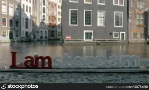 View of small plastic figure of Iamsterdam letters sculpture on the bridge against blurred cityscape with building on the embankment of canal and water of river, Amsterdam, Netherlands