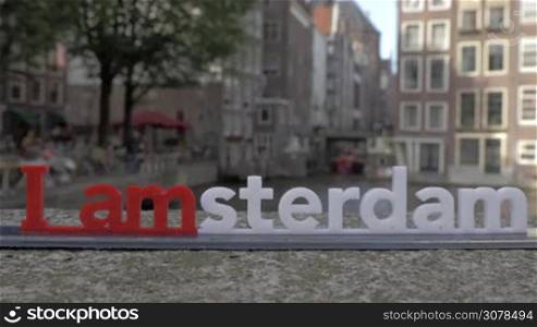 View of small plastic figure of Iamsterdam letters sculpture on the bridge against blurred cityscape with building on the embankment of canal, Amsterdam, Netherlands