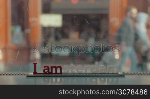 View of small plastic figure of Iamsterdam letters sculpture on the scratched windows on tram against blurred cityscape with buildings and road and bikes, Amsterdam, Netherlands