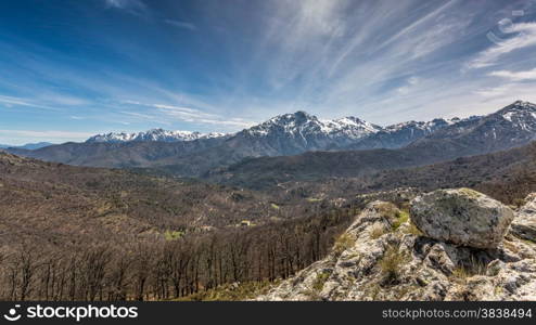 View of snow covered Monte Pardu and San Parteo in the Balagne region of north Corsica with rocks, trees and maquis in the foreground and blue skies and wispy clouds