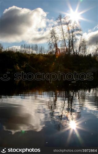 View on autumn landscape of river and trees in evening. Forest on river coast in autumn evening. Reflection of autumn trees, clouds and sun in water. Autumn in Latvia. Autumn landscape with colorful trees and river. Evening reflection in river.