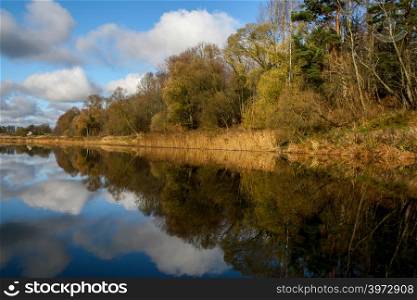 View on autumn landscape of river and trees in sunny day. Forest on river coast in autumn day. Reflection of autumn trees and clouds in water. Autumn in Latvia. Autumn landscape with colorful trees, yellow grass and river.