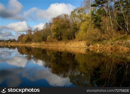 View on autumn landscape of river and trees in sunny day. Forest on river coast in autumn day. Reflection of autumn trees and clouds in water. Autumn in Latvia. Autumn landscape with colorful trees, yellow grass and river. 