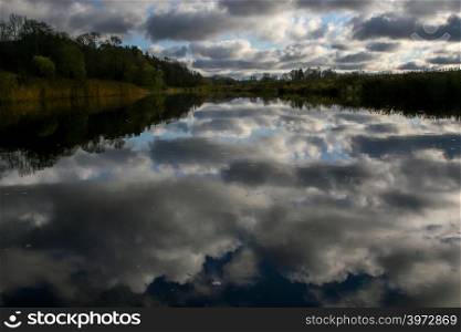 View on summer landscape of river and trees in evening. Forest on river coast in summer day. Reflection of clouds and sky in water. Summer in Latvia. Autumn landscape with colorful trees, grass and river.