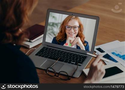 View over shoulder of female freelancer talks with employee by video call, discuss strategy of work. Laptop screen view of smiling ginger woman in spectacles. New technology usage. Distance working