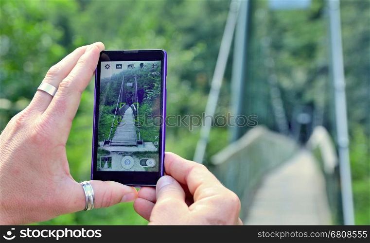 View over the mobile phone display during taking a picture of bridge in nature. Man is holding the mobile phone in hand and taking a photo in nature. Closeup view on hands with mobile phone.