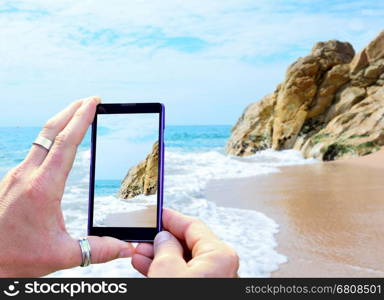 View over the mobile phone display during taking a picture of Costa Brava beach in Calella, Spain. Holding the mobile phone in hands and taking a photo. Focused on mobile phone screen.