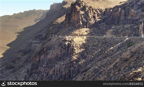 View over the rugged rocks of the mountains on the canary island of Gran Canaria in the afternoon sun. A road leads on the steep mountain down into the valley.