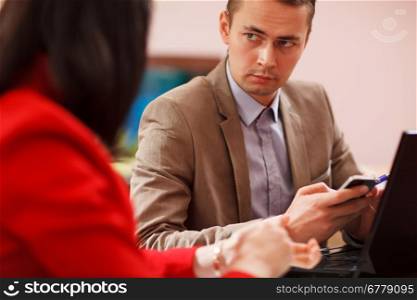 View past the shoulder of a woman to an earnest young businessman listening to a colleague as he sits at his desk with his mobile in his hand