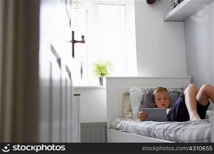 View through doorway of boy lying on bed looking down at digital tablet