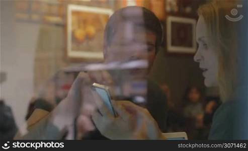 View through restaurant glass of woman and man talking cheerfully, woman shows something on the smartphone