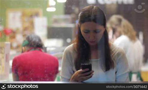 View through restaurant glass of young brunette woman typing using smartphone while waiting for her friend, she smiles and lifts her head up to look at something