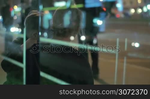 View through the glass of a mother with son on the laps sitting and waiting at the outdoor bus stop in night city