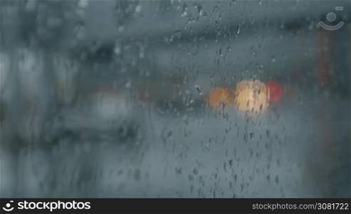 View through wet glass to the car with headlights driving on the road. Focus on the window with raindrops