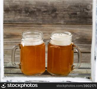 View through windows of two glass jars filled with frosty cold beer on top of wooden crate with rustic wood in background.