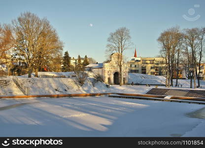 view to the gate of Tallinn in Parnu, Estonia with a bandstand surrounded by trees in foreground on sunny winter day, with the moon over it in the clear blue sky