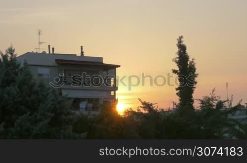 View to the house with balconies with green trees and bushes in foreground, golden sunset being seen from behind the building