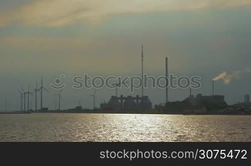 View to the industrial area of the city located on the shore. Working windmills and factories. Water sparkling in the sun in foreground
