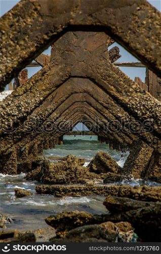 View under at Broken old structure remains of pier in the sea. Small wave crashing into the textured rusty pier posts.