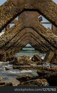 View under at Broken old structure remains of pier in the sea. Small wave crashing into the textured rusty pier posts.