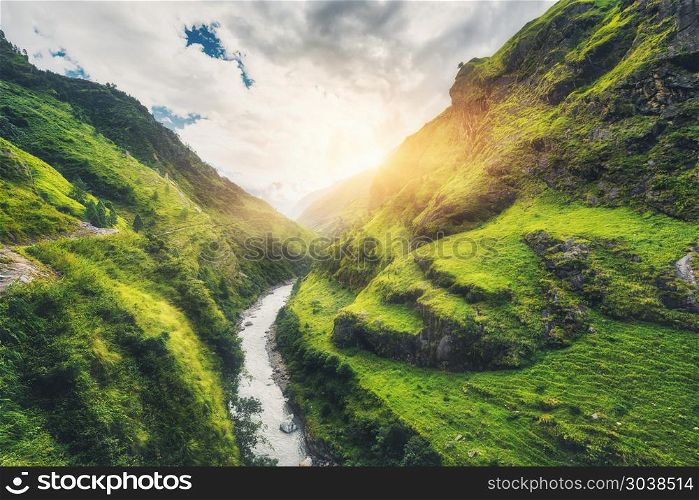 View with amazing mountains covered green grass, river, meadows and forest, blue sky with clouds, sun in autumn in Nepal at sunset. Mountain valley at bright sunny evening. Travel in Himalayas. Nature. Amazing mountains covered green grass, river at sunset