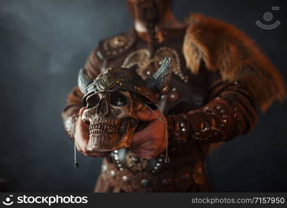 Viking dressed in traditional nordic clothes holds human skull in helmet, barbarian image. Ancient warrior in smoke on dark background. Viking holds human skull in helmet