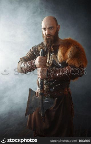 Viking with axe dressed in traditional clothes, nordic barbarian image. Ancient warrior in smoke on dark background