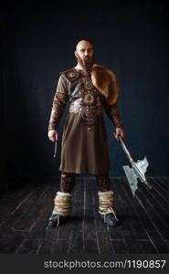 Viking with axe dressed in traditional nordic clothes. Ancient warrior, studio with black walls and wooden floor on background