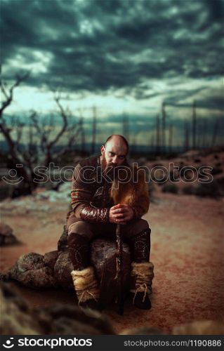 Viking with axe dressed in traditional nordic clothes sitting on stone in wasteland, battle in rocky mountains. Scandinavian ancient warrior