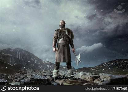 Viking with axe dressed in traditional nordic clothes standing in rocky mountains. Scandinavian ancient warrior