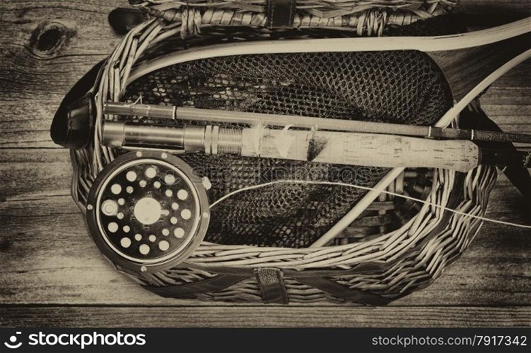 Vintage and aging grain concept of an antique fly fishing reel, rod, flies, and net on top of open creel with rustic wood underneath. Layout in horizontal format.