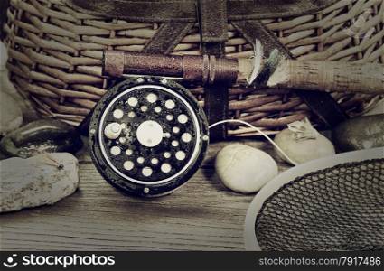 Vintage concept with grain of a wet antique fly fishing reel, rod, landing net, artificial flies and rocks in front of creel with rustic wood underneath. Layout in horizontal format.
