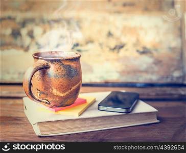 Vintage cup with hot drinks and steam, tee or coffee on old book with smart-phone on dark rustic wooden table