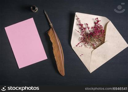 Vintage envelope full with pink flowers, an antique quill pen, an ink pot and a blank message card, on a black wooden background.