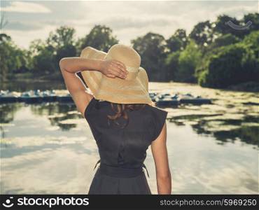 Vintage filtered shot of an elegant woman wearing a hat standing by a lake in a park at sunset