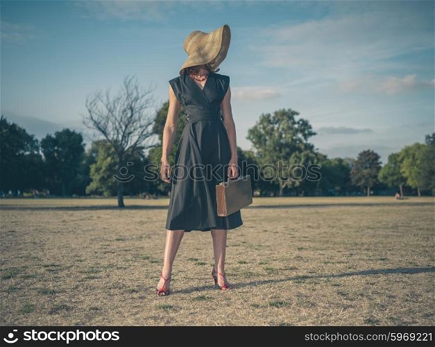 Vintage filtered shot of an elegant young woman wearing a dress and a hat standing in the park with a briefcase