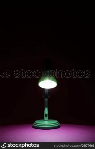 Vintage green desk lamp in the dark turned on and shining on a bright pink work surface. Copy space and room for text on a vertical orientation image.