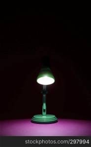 Vintage green desk lamp in the dark turned on and shining on a bright pink work surface. Copy space and room for text on a vertical orientation image.