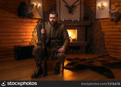 Vintage hunter man in traditional hunting clothing sitting in a chair with retro rifle against burning fireplace. Stuffed wild animals, bear skin and other trophies on background. Vintage hunter man in traditional hunting clothing