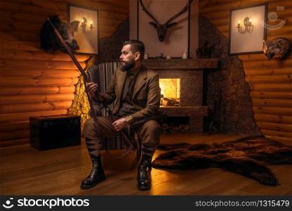 Vintage hunter man in traditional hunting clothing sitting in a chair with retro rifle against burning fireplace. Stuffed wild animals, bear skin and other trophies on background