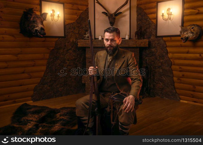Vintage hunter man in traditional hunting clothing sitting in a chair with retro rifle against burning fireplace. Stuffed wild animals, bear skin and other trophies on background. Vintage hunter man in traditional hunting clothing