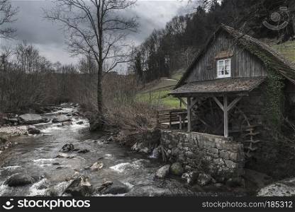Vintage landscape with an old, abandoned water mill on an alpine river shore, on a cloudy day of early spring, in Schwarzwald, Germany.