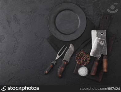 Vintage meat knife and fork and hatchets with vintage chopping board and plate on black table background. Butcher utensils.
