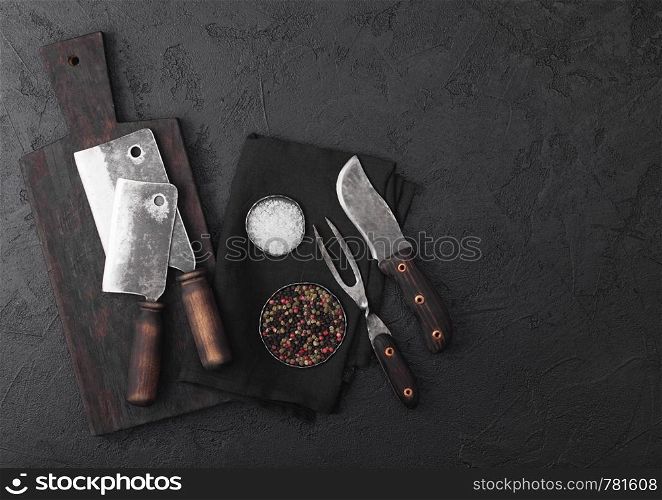 Vintage meat knife and fork and hatchets with vintage chopping board and black table background. Butcher utensils.