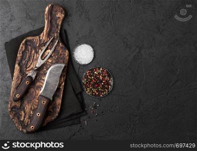 Vintage meat knife and fork on vintage chopping board and black stone table background. Butcher utensils.