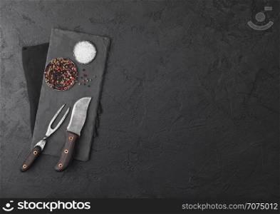 Vintage meat knife and fork with stone chopping board and black table background. Butcher utensils.