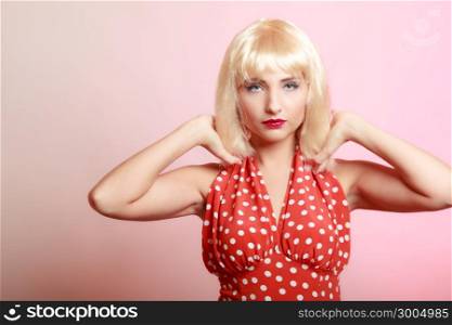 Vintage pinup style. Portrait of beautiful stylized young woman. Attractive girl in blond wig and retro spotted red dress on pink. Disguise. Studio shot.