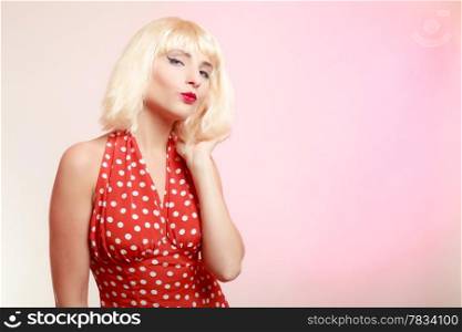 Vintage pinup style. Portrait of beautiful stylized young woman flirting blowing a kiss. Attractive girl in blond wig and retro spotted red dress on pink. Disguise. Studio shot.