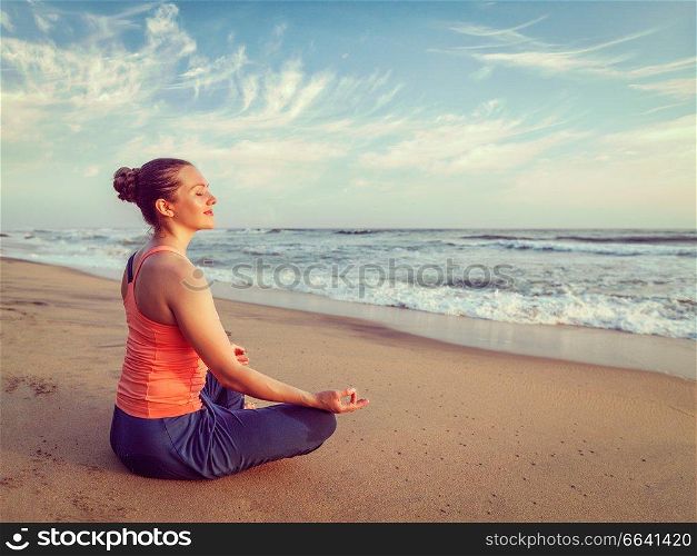 Vintage retro hipster effect image of woman doing yoga meditating and relaxing in Padmasana Lotus Pose with chin mudra outdoors at tropical beach on sunset. Young sporty fit woman doing yoga oudoors at beach